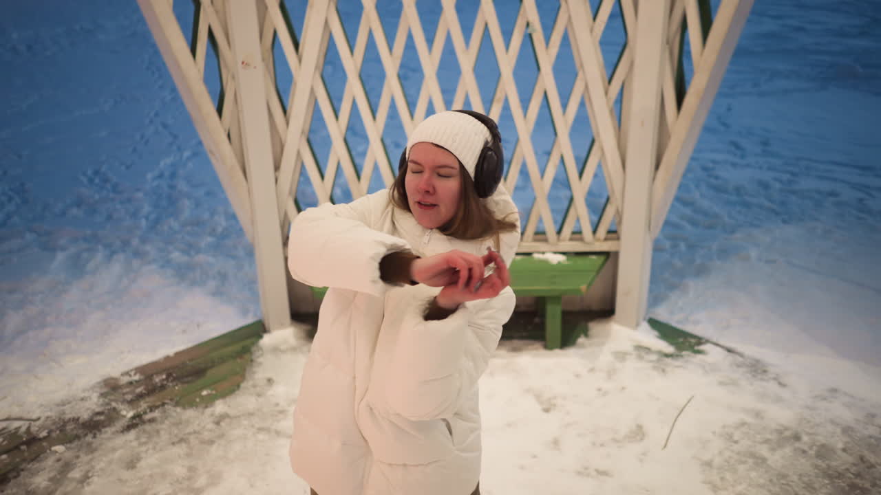 Snow dancer moves with closed eyes wearing headphones in illuminated gazebo draped in lattice panels, wearing white puffer coat and beanie as gentle evening breeze swirls around snowy ground