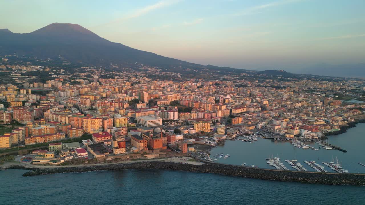 A breathtaking drone shot showing an Italian harbour town at sunrise, with sunlit coastal houses a peaceful harbour, and majestic mountains in the backdrop highlighting the beauty of the Mediterranean