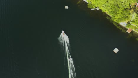 at midday, a man is seen water skiing or wakeboarding rapidly on a lake with many lush, green trees in the backdrop