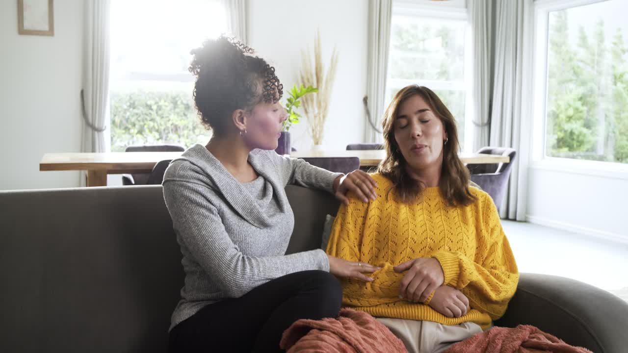 Diverse Freundinnen umarmen sich auf dem Sofa neben einer Pflanze zu Hause, trösten eine verärgerte Freundin und ermutigen sie zum Reden.