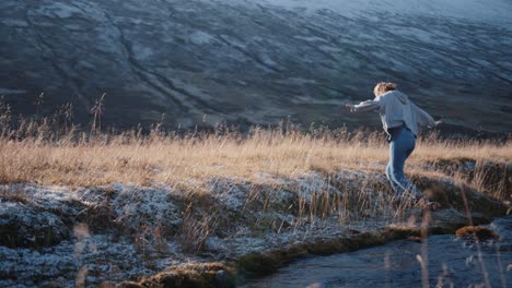 Woman-running-and-jumping-above-river-in-bright-day-near-snowy-mountains