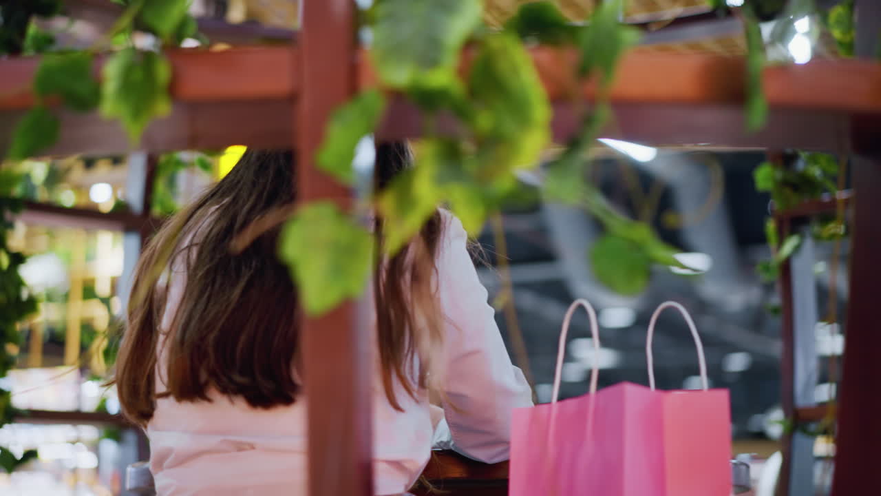 양쪽에 쇼핑 가방이 있는 잘 꾸며진 술집에 앉아 있는 여성의 뒷면, 배경에 활기찬 장식과 함께 활기찬, 세련된 분위기를 포착합니다.