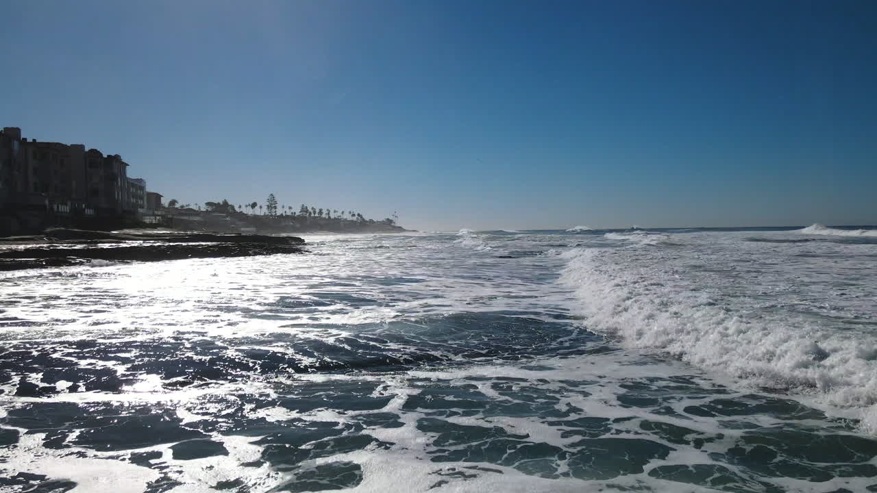 A low-flying drone glides just above the waves of the beach, capturing the dynamic movement of the water and the shimmering reflections of sunlight.