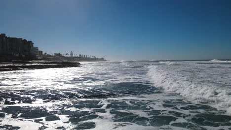 a low-flying drone glides just above the waves of the beach, capturing the dynamic movement of the water and the shimmering reflections of sunlight.