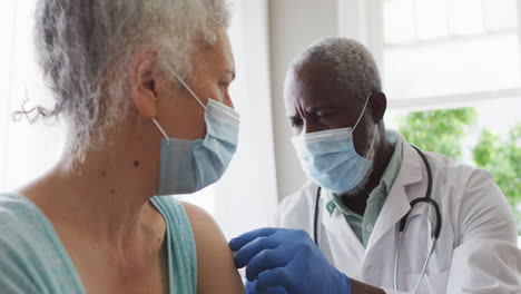 male african american doctor wearing face mask injecting covid-19 vaccine into senior female patient