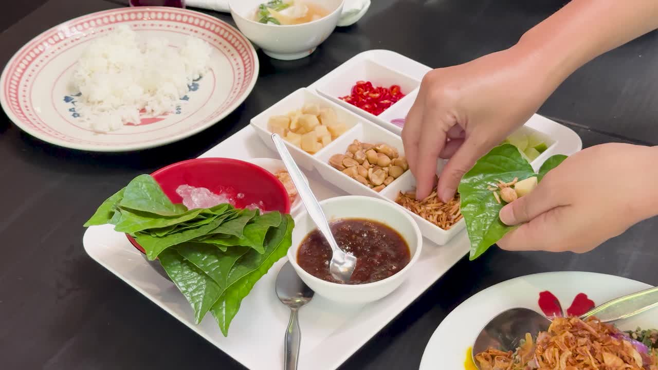 Hands prepare traditional Miang Kham by adding peanuts, dried shrimp, ginger, and sauce to wild betel leaves on a well-lit dining table