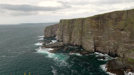 Seabirds-fly-above-a-turquoise-green-ocean-in-front-of-a-steep-and-imposing-sea-cliff-rising-straight-up-out-of-the-ocean-as-waves-crash-against-its-base