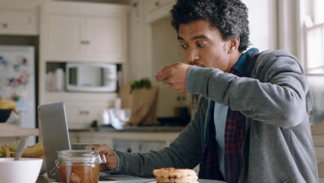 happy mixed race man using laptop computer working in kitchen eating waffles for breakfast browsing online enjoying relaxing morning at home