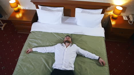 tired businessman on comfortable bed. man in white shirt and trousers laying on bed relaxing in hotel after the hard journey. top view.