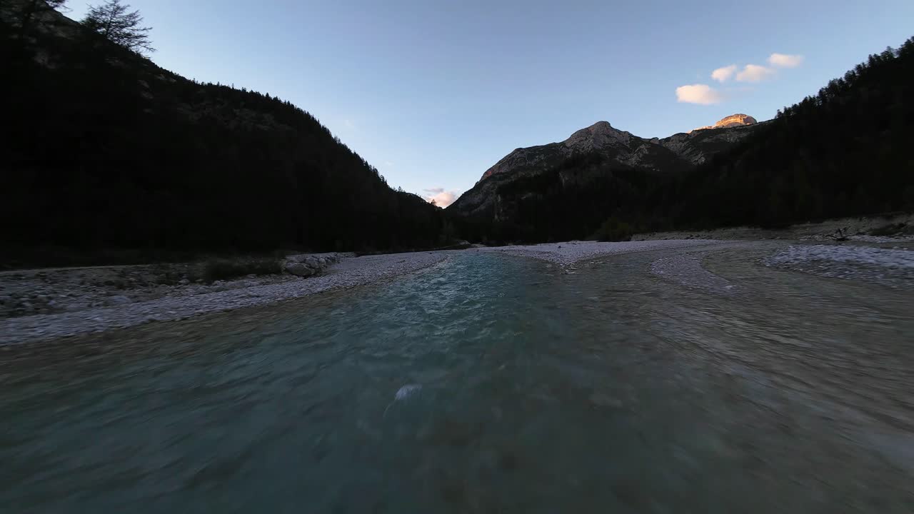 Drone dengan sudut pandang orang pertama terbang rendah dan cepat di atas sungai pegunungan yang jernih, melaju melewati lembah yang menakjubkan saat sinar matahari terbenam terakhir menerangi puncak-puncak megah di latar belakang.