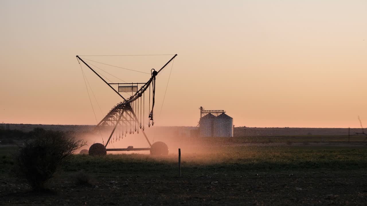 Backlit golden hour irrigation pivot waters crop field, orange sky