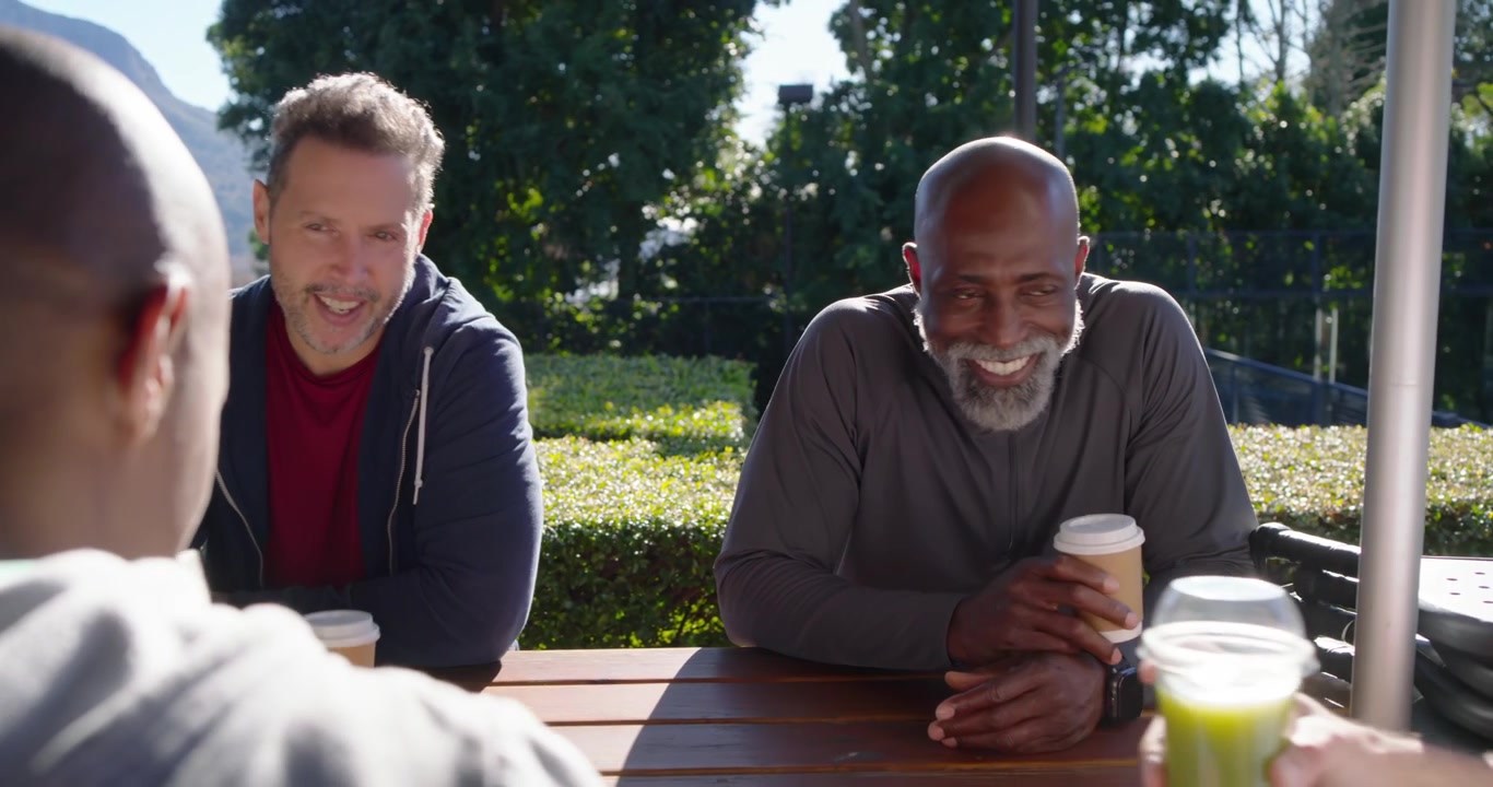 Diverse male friends reaching in, clinking paper cups and plastic cups at picnic table, catching up