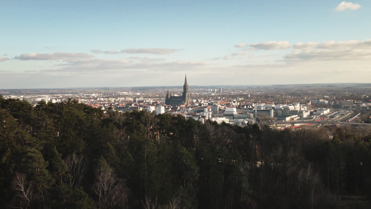 Drone aereo 4K che si alza attraverso la foresta rivelando il paesaggio urbano di Ulm e il punto di riferimento della cattedrale. Filmato Premium che cattura la chioma degli alberi, lo skyline della città e il paesaggio panoramico. Perfetto per documentari sulla natura e sui viaggi.