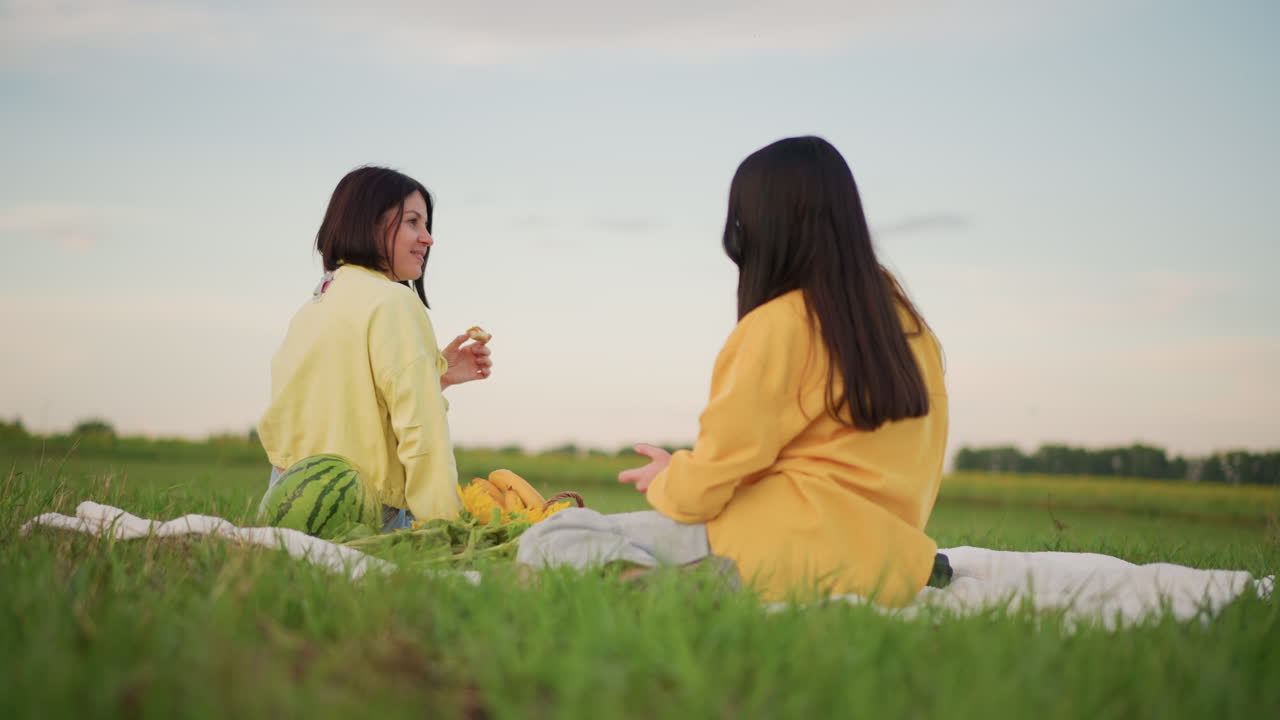 황혼녘 탁 트인 들판에서 담요를 덮고 앉아 간식과 시원한 음료를 나눠 마시며, 아늑한 노란색 스웨터를 입고 들꽃으로 둘러싸인 채 웃고 있는 두 백인 여성. 피크닉 바구니가 보이고 편안하고 장난기 넘치는 분위기의 자연스러운 모습