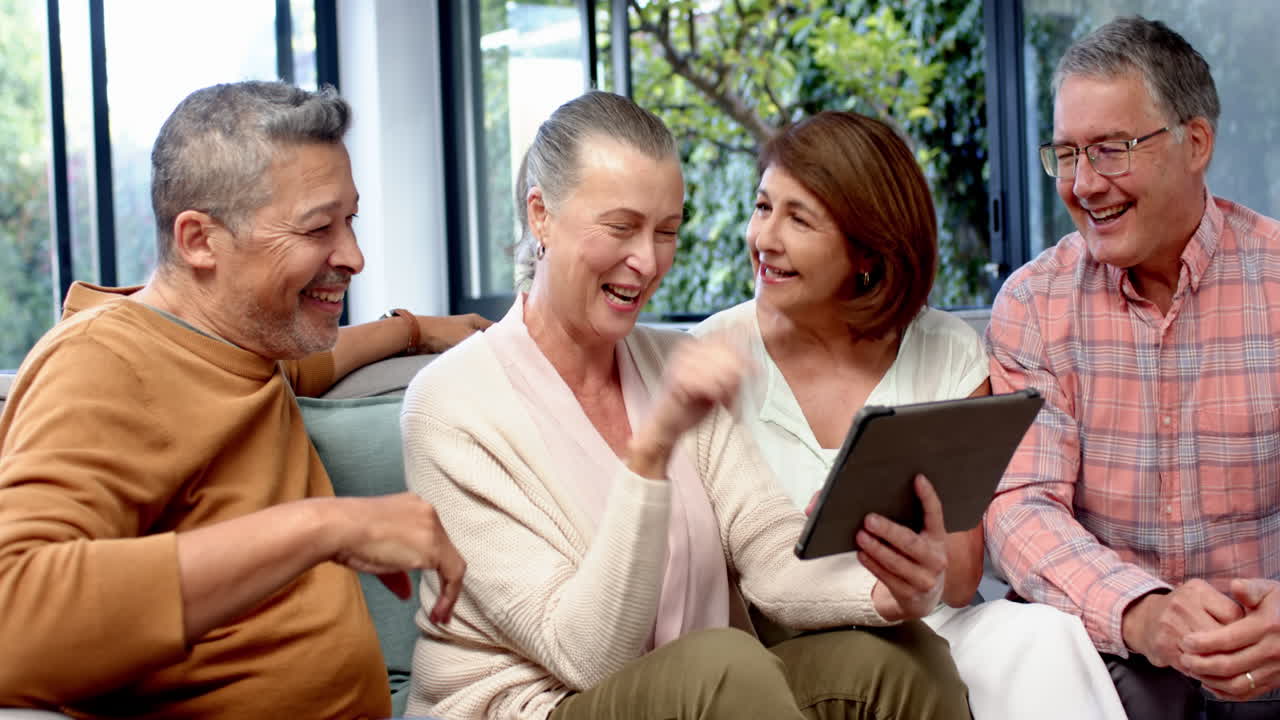 Seniors sitting on couch, using tablet and laughing together, enjoying time