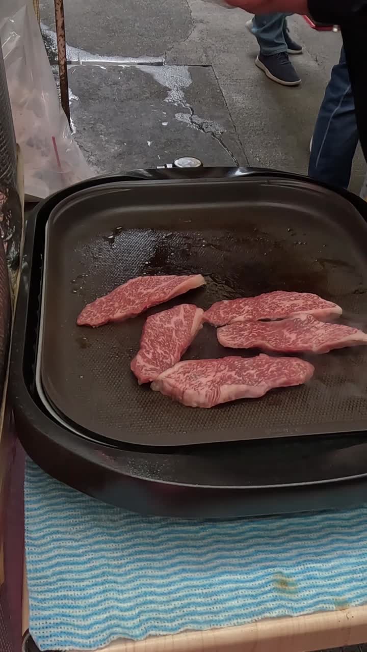 Close-up irisan daging sapi marmer premium mendesis di atas wajan hitam di pasar jalanan.