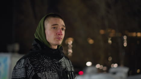 side view of man wearing green hoodie and patterned winter jacket walking outdoors at night in cold weather with visible breath escaping mouth, blurred bare trees and soft bokeh lights in background