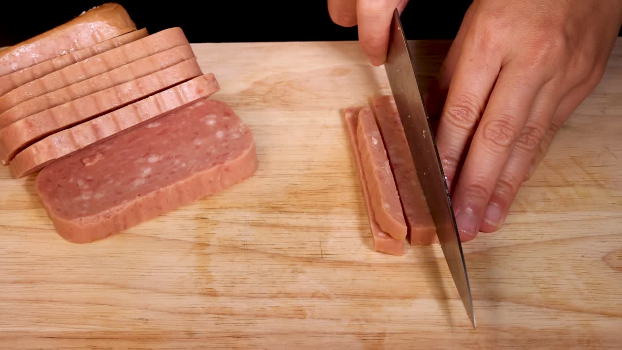 A person uses a kitchen knife to slice processed luncheon meat into strips on a wooden cutting board under even, bright lighting with a top-down view