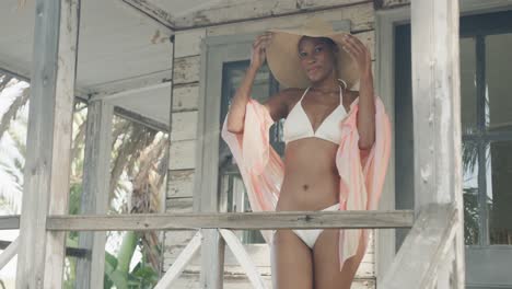 retrato de mujer biracial en bikini y sombrero de sol sonriendo en el porche de una casa de playa de madera, cámara lenta