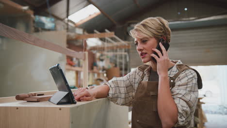 female apprentice carpenter with digital tablet working in furniture workshop making phone call