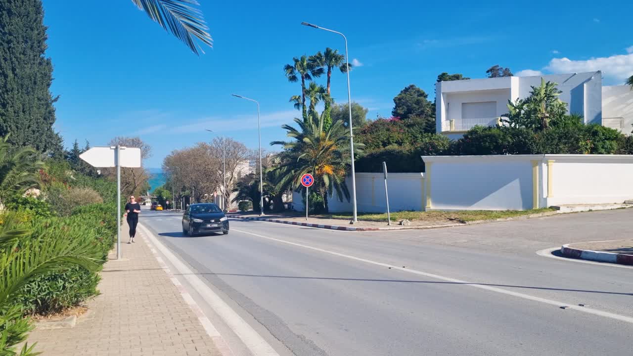 Cars, motorcyclist, and female jogger on a palm lined road with white buildings under a blue sky. Filmed on a sunny spring day in Sidi Bou Said close to Tunis, Tunesia