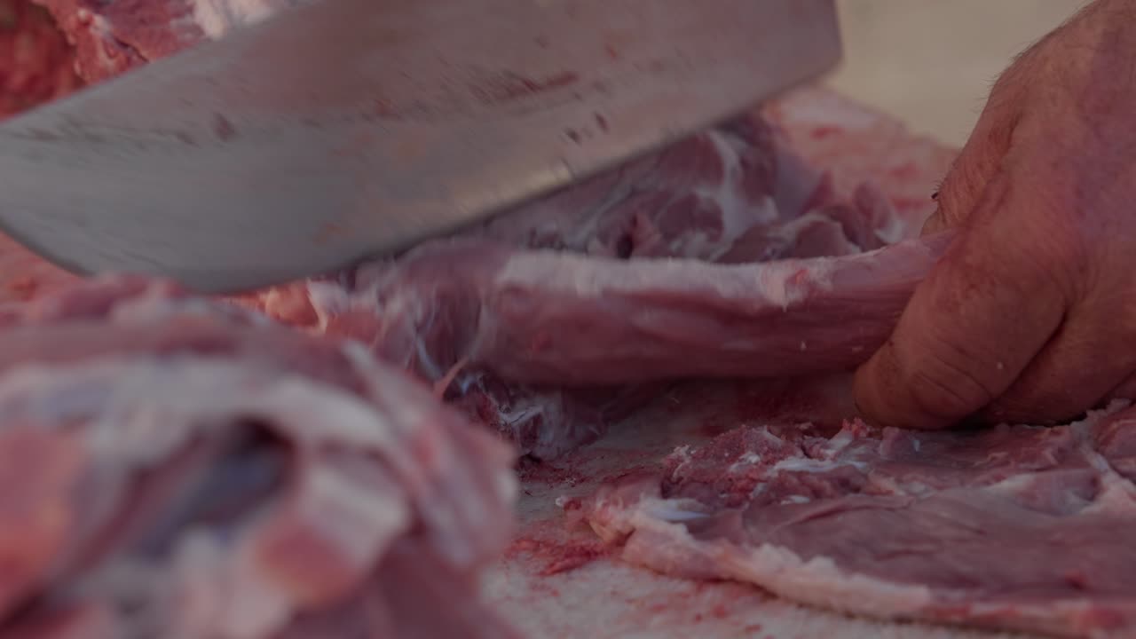 hand holds pork slab while cleaver cuts meat on bloodstained surface