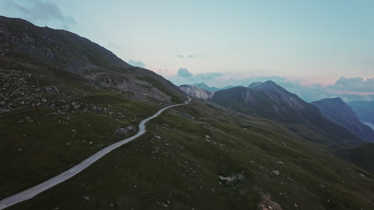 En snoet vej gennem grønne bjerge ved solnedgang med fjerne bjergtoppe i syne, luftfoto.