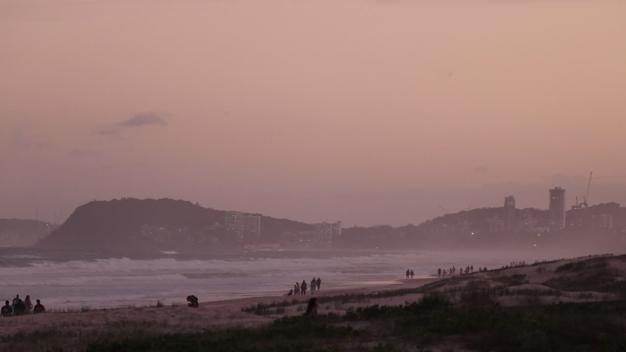gente paseando por la playa al anochecer, la ciudad en el fondo