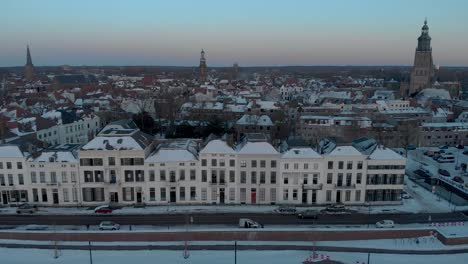 Cityscape-sunset-aerial-pan-of-Dutch-Hanseatic-medieval-tower-town-Zutphen,-The-Netherlands,-with-snow-on-the-boulevard-lit-up-by-last-daylight-with-river-IJssel-in-the-foreground