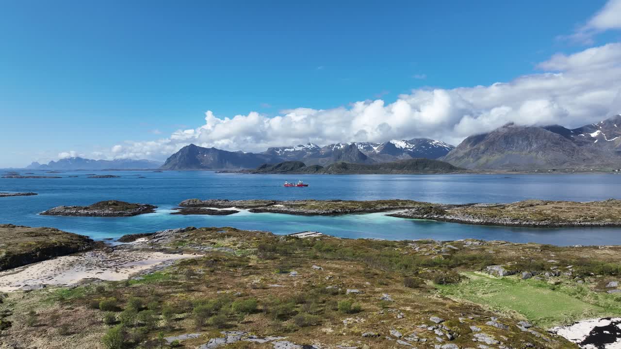 Low aerial flying toward aquaculture well boat sailing through Gimsoy current, Lofoten, Norway with peaks, beach of Lofoton in background