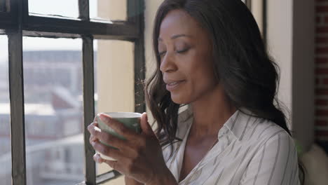 Hermosa-Mujer-Afroamericana-Tomando-Café-En-Casa-Disfrutando-De-Un-Estilo-De-Vida-Exitoso-Mirando-Por-La-Ventana-Planeando-Con-Anticipación-Una-Mujer-Negra-Madura-Relajándose-En-Un-Acogedor-Apartamento-Tipo-Loft