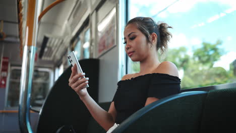 Una-Joven-Asiática-Vestida-Con-Un-Top-Negro-Con-Hombros-Descubiertos,-Se-Concentra-Intensamente-En-Su-Teléfono-Inteligente-Mientras-Está-Sentada-En-La-Ventana-De-Un-Tren,-Resalta-Su-Rostro-Y-El-Moderno-Interior-Del-Tren.