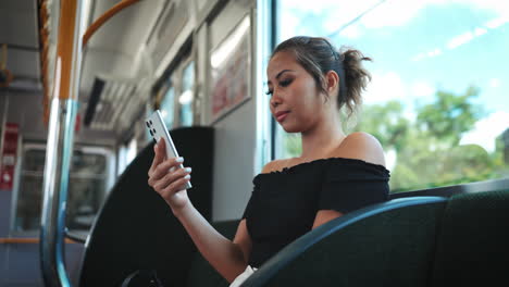 young asian woman wearing a black off-shoulder top, focuses intently on her smartphone while seated on a train window highlights her face and the modern train interior