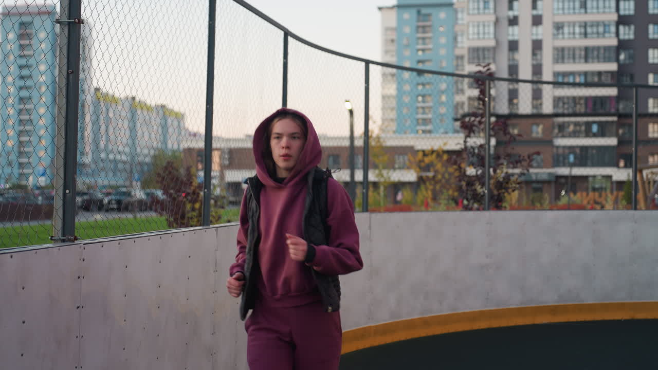 Fitness enthusiast jogging on circular urban sports court with white post under twilight sky against apartment towers, steady pace warmup in hoodie vest and jogger pants with white sneakers