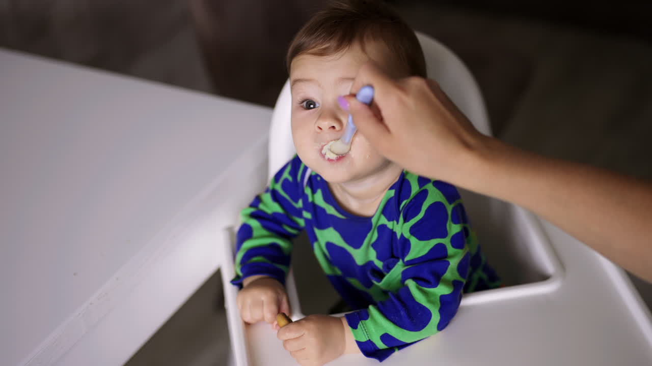 Lovely boy in blue shirt eats dairy from spoon. Cute baby sitting in white feeding chair looks aside. High angle view.