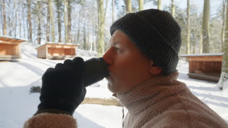 close-up of a person in winter attire drinking from a thermos outside snowy cabins