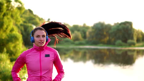 morena con cabello largo en auriculares corre a lo largo del río en el parque por la mañana al amanecer en el verano en una chaqueta rosa y pantalones negros
