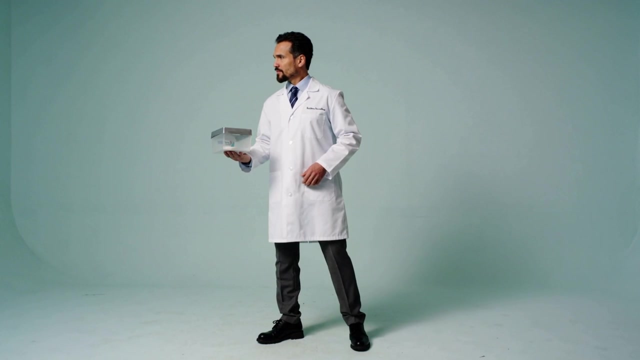 Entering from right, man in lab coat and tie walking into mint-green studio, presenting clear box
