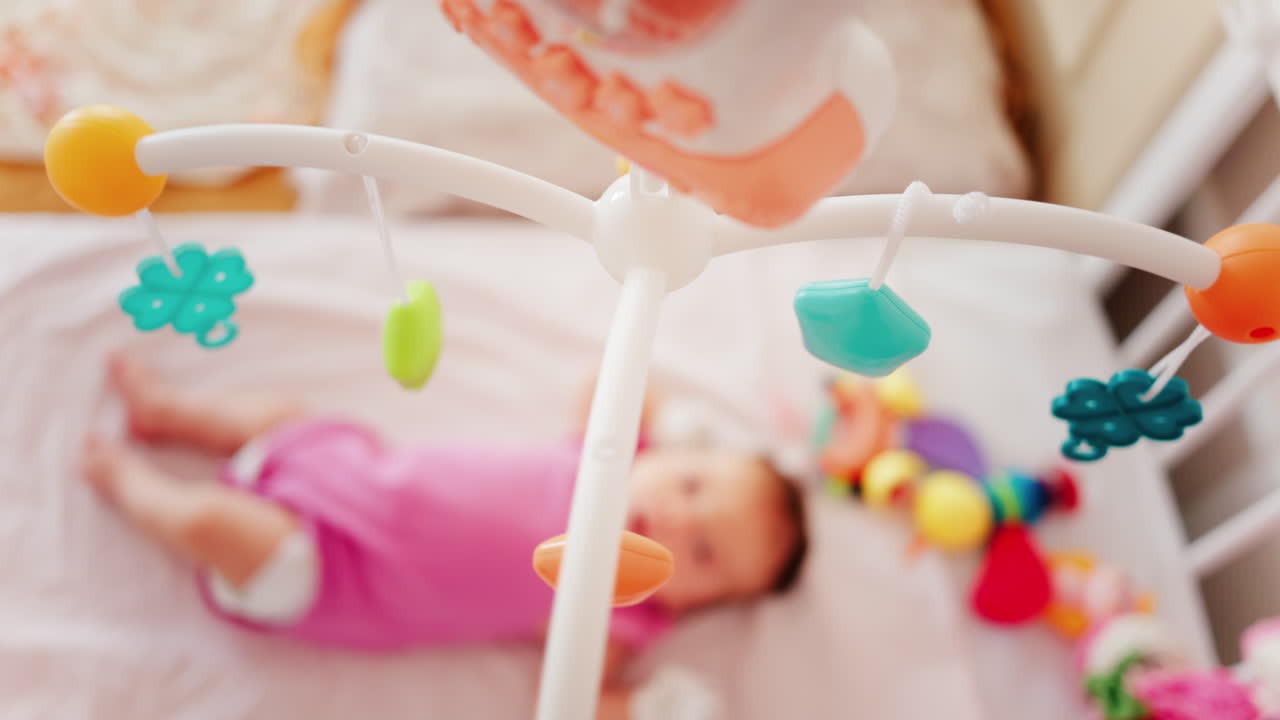 Baby lying on a bed in a pink outfit, looking up at a colourful hanging mobile toy
