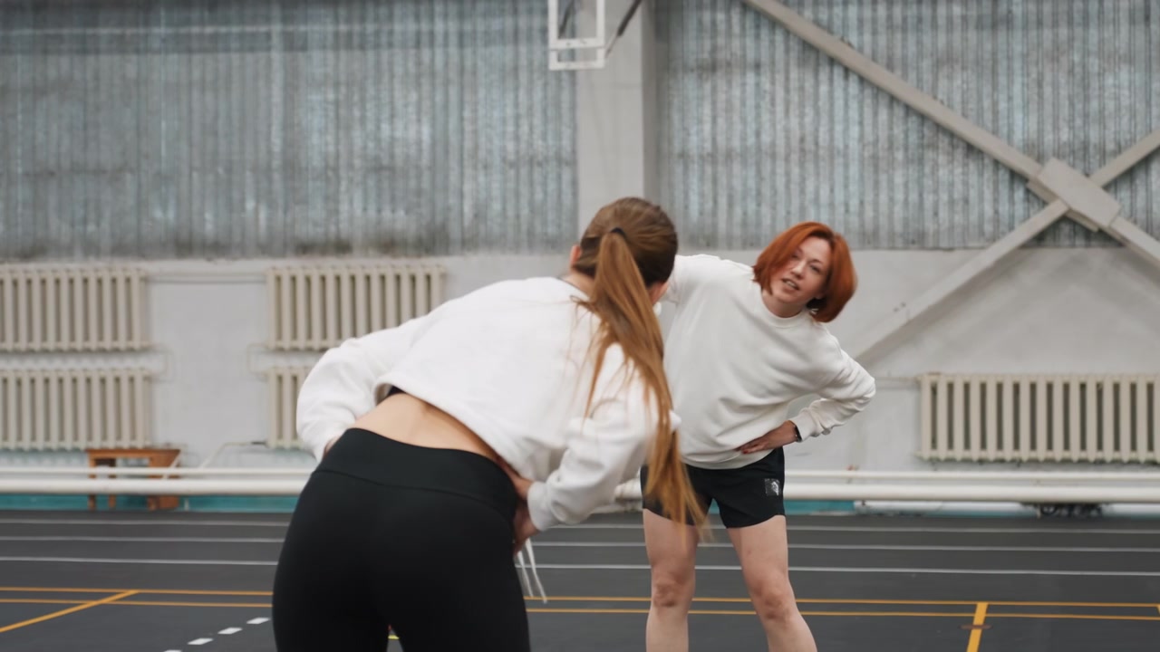 White women badminton partner warmup on court, playful exchange and mirrored stretches, hands on hips and forward bends, short shorts and sweatshirt contrast, indoor training energy and teamwork
