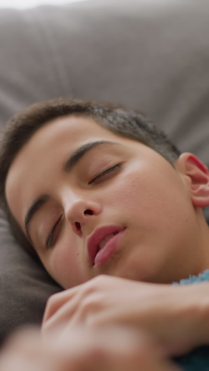 Close-up of sick child resting on bed covered with blue blanket, turning with discomfort while coughing, with tissue nearby and light streaming through window