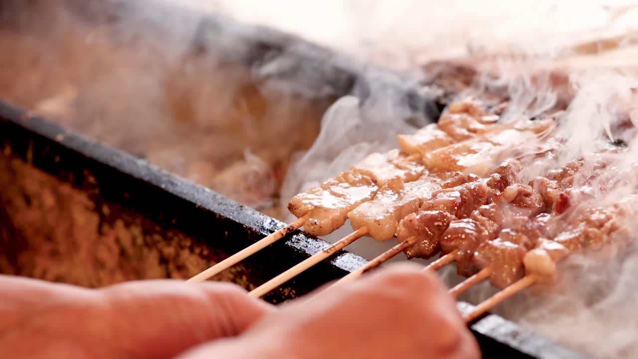 Close-up of hands turning skewered meat over a smoky grill, highlighting the cooking process and smoke.