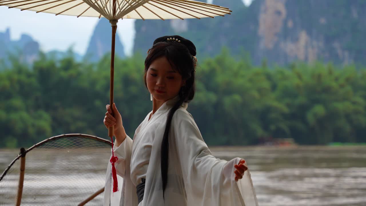 Beautiful Hanfu girl with umbrella posing on a bamboo raft extending arms and fixing her hair