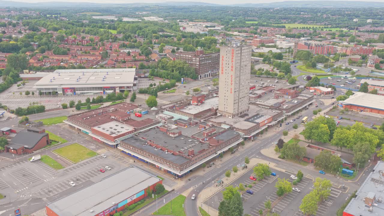 The Salford Shopping City complex, anchored by its tall residential tower, is surrounded by retail units, car parks, and suburban neighborhoods, captured in a descending aerial drone shot