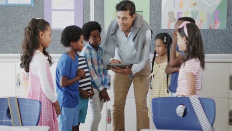happy diverse male teacher with schoolchildren using tablet in classroom at elementary school