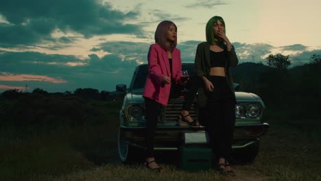two women relaxing by a vintage car at sunset
