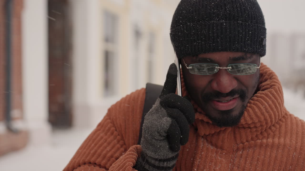 Black man walking snowy street on phone, smiling and talking, wearing orange ribbed sweater, black beanie and clear glasses backpack over shoulder while passing colorful storefronts and brick facades