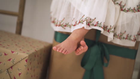 Slow-motion-of-Cute-Girl-dressed-in-festive-dress-sitting-on-Christmas-Gifts-swinging-her-feet---close-up