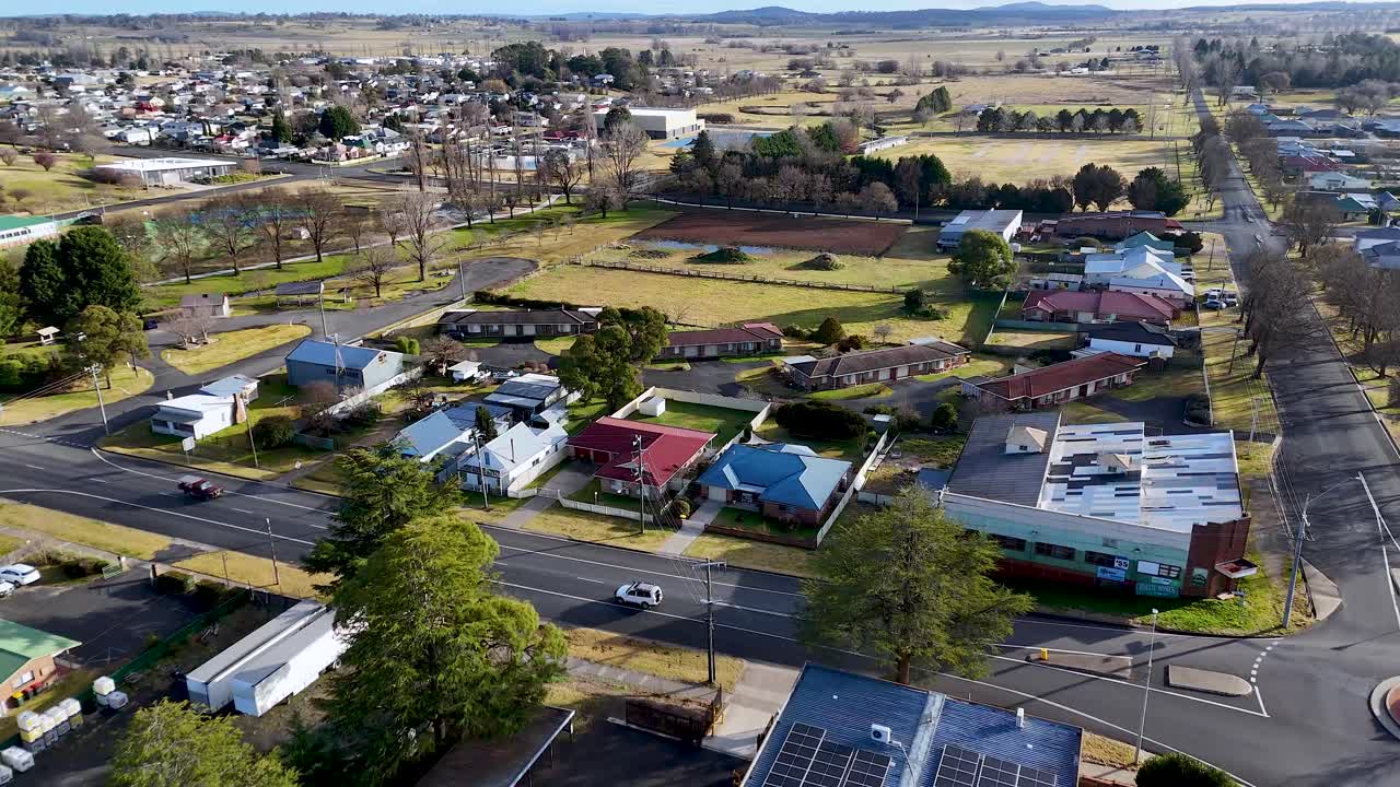 Drone camera glides above a quiet suburban intersection in Glen Innes, Australia, revealing residential homes, commercial buildings, and tree-lined streets in clear daylight