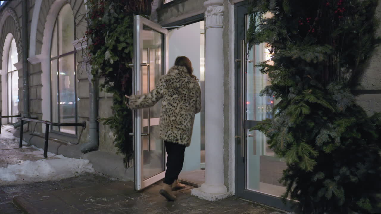 Stylish woman in spotted winter jacket approaches glass door entrance surrounded by evergreen decorations on snowy evening. City lights and icy sidewalk create cozy cold weather atmosphere